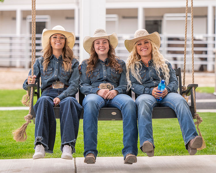 Three cowgirls smiling at the camera, sitting on a porch swing wearing denim shirts.
